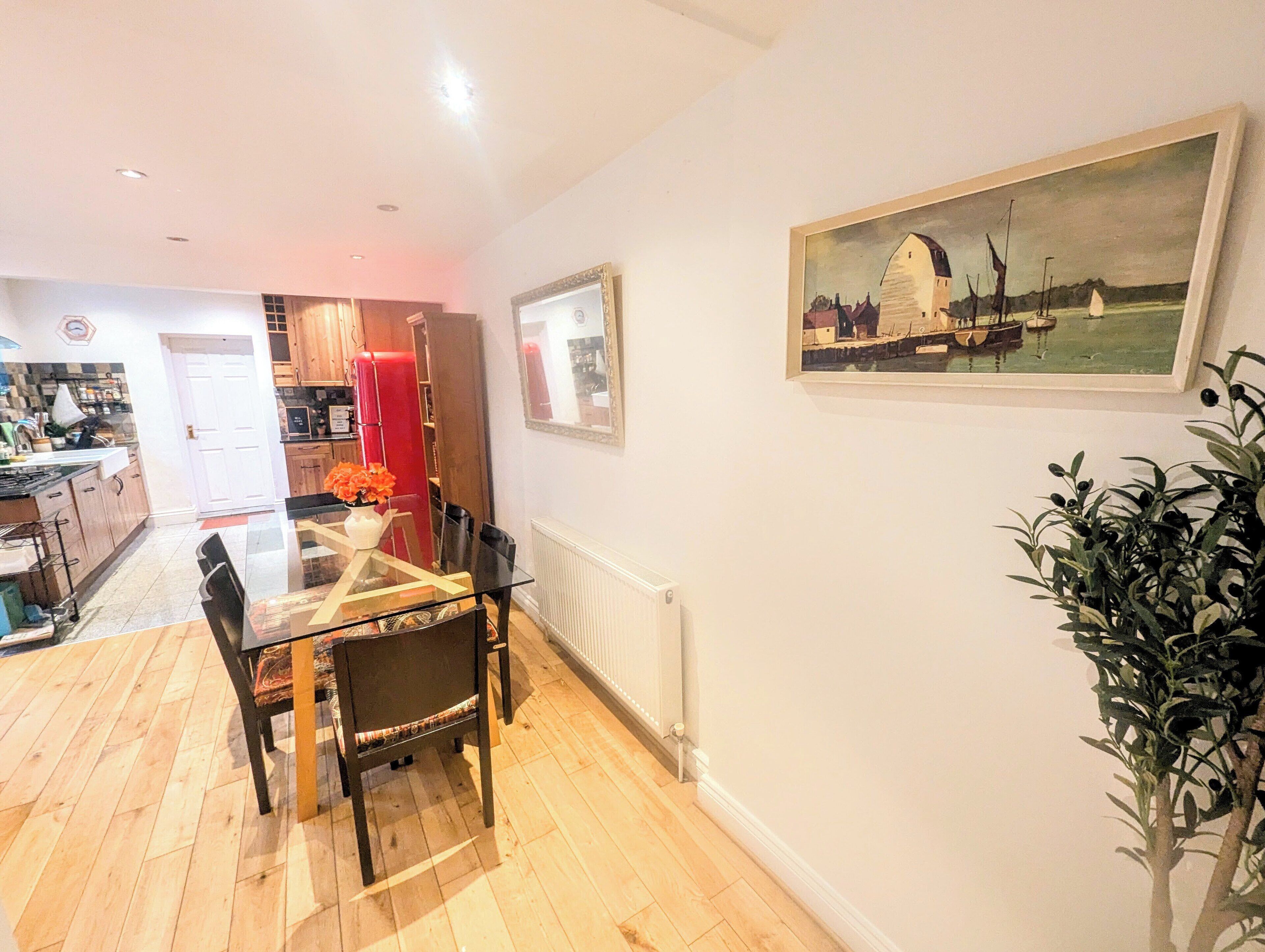 A dining area with a glass table and chairs, a red curtain, and a painting of a coastal scene.