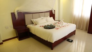 A neatly made bed with white linens, a decorative swan pillow, and rose petals. There is a wooden nightstand with a lamp, and a pair of slippers on the floor.