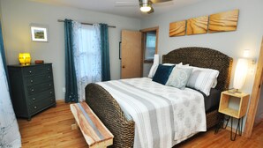 Un dormitorio con cama, un banco de madera, cómoda, mesita de noche, ventilador de techo y ventana con cortinas.