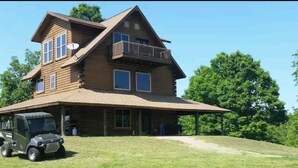 A two-story log cabin with a large porch, a satellite dish, and a vehicle parked in front. - LOG HOME CANINE RETREAT (Pyatt)