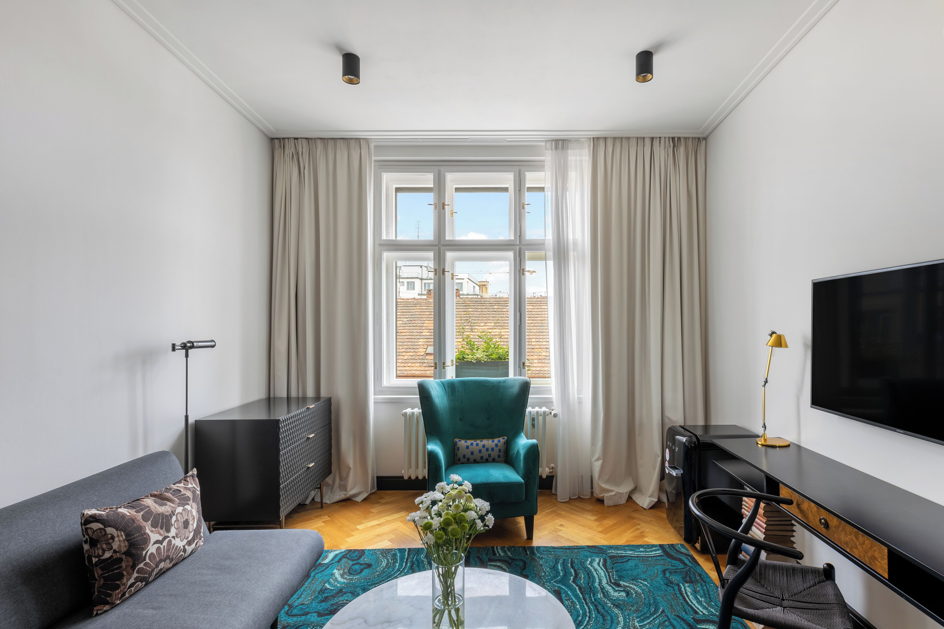 Een moderne woonkamer met een bank, een blauwgroene fauteuil, een salontafel met bloemen, een televisie en een bureau met een lamp.