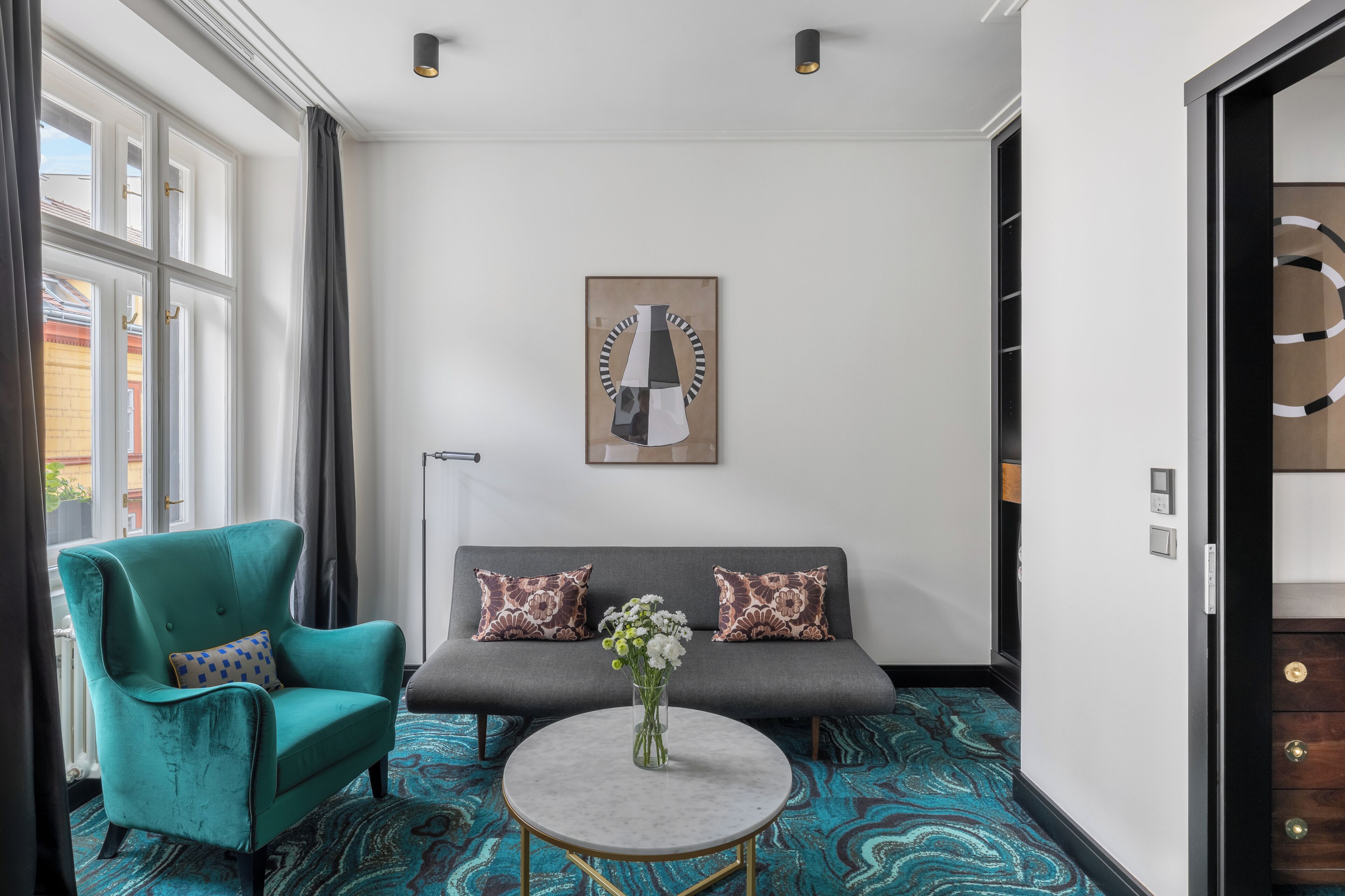 Ein modernes Zimmer mit einem türkisfarbenen Sessel, einem grauen Sofa mit gemusterten Kissen, einem runden Tisch mit Blumenvase und einem großen abstrakten Gemälde an der Wand.