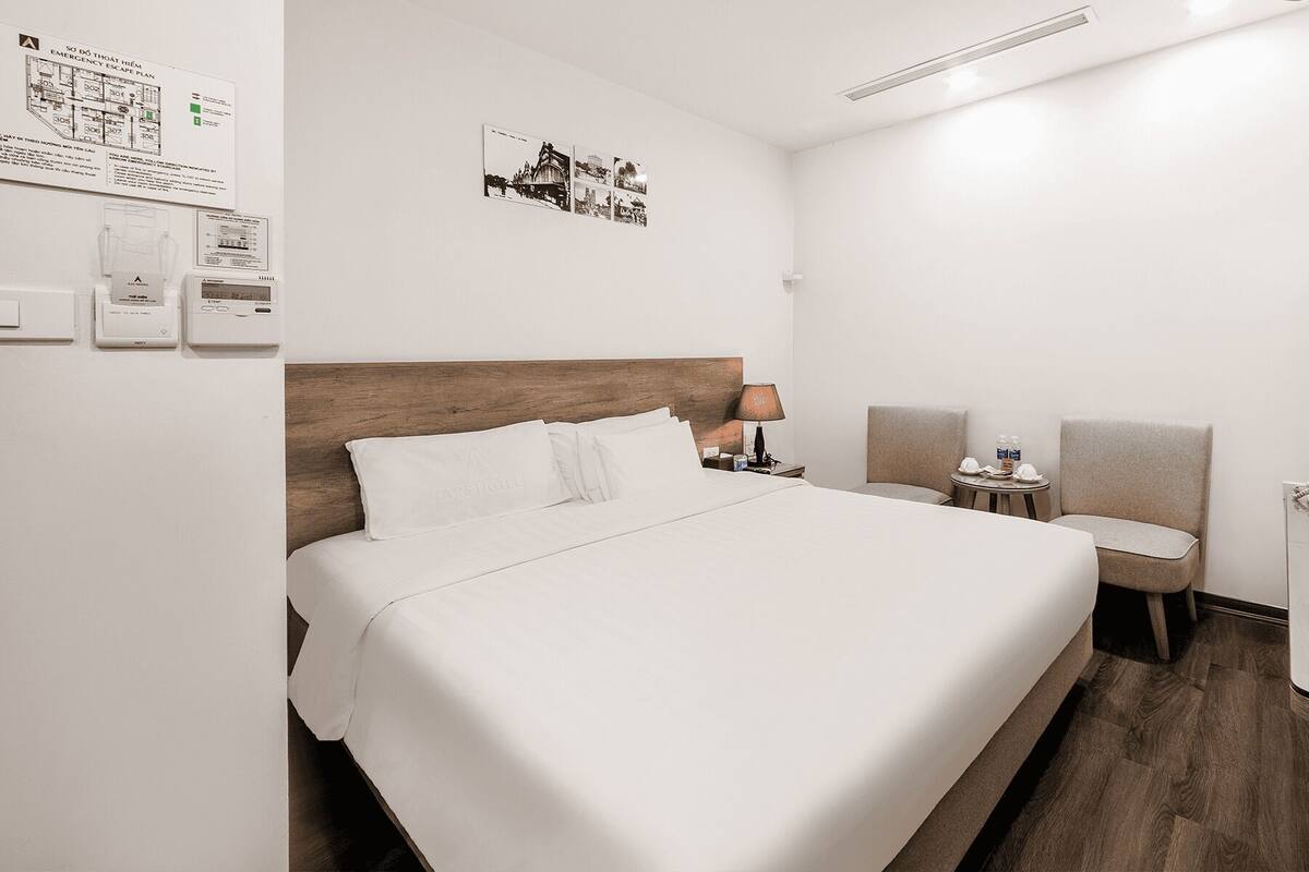A hotel room with a large bed, a bedside table with a lamp, a chair, a small table with a water bottle, and a phone.