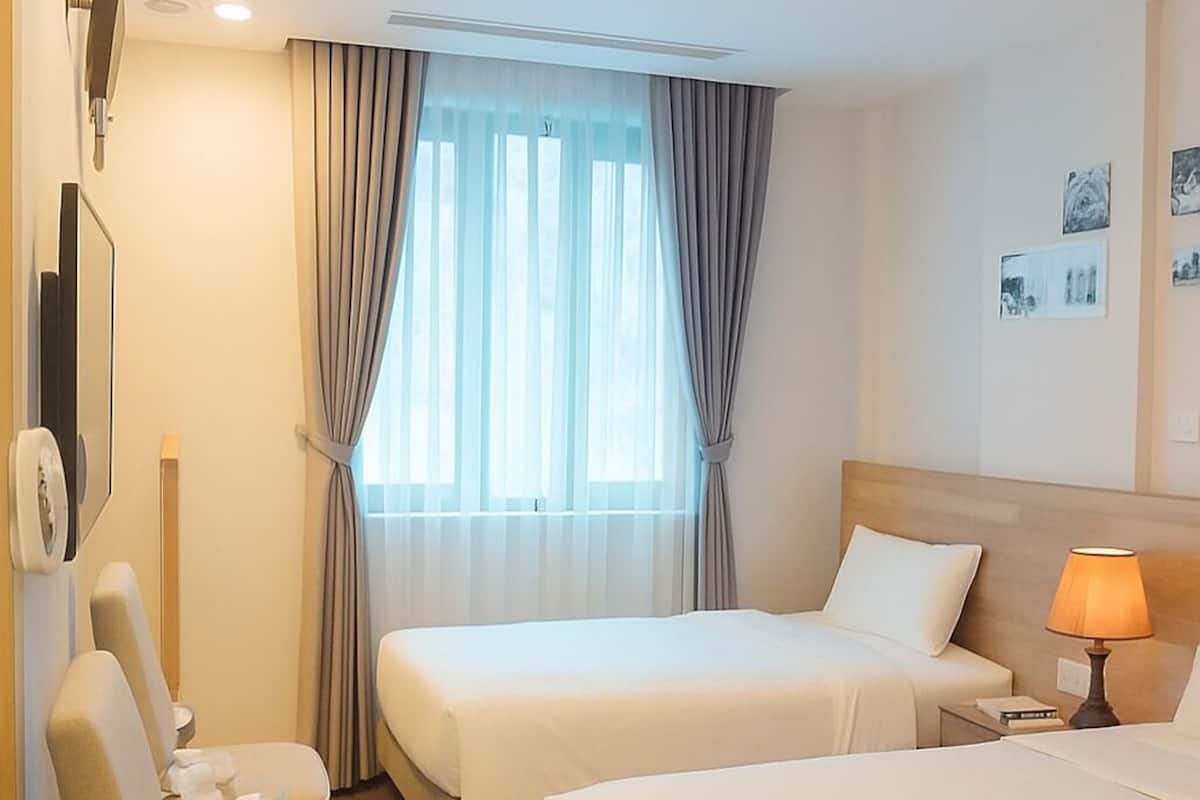 A hotel room with two beds, a chair, a bedside table with a lamp, and a window with curtains.