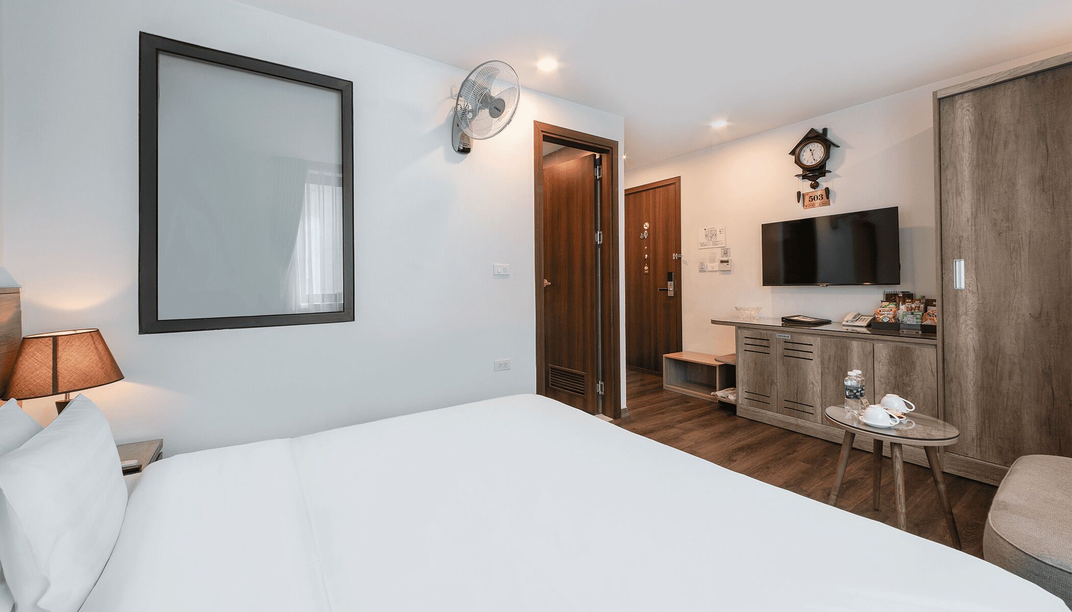 Habitación de hotel con cama, escritorio con televisor de pantalla plana, armario de madera, reloj y espejo.