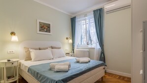 Ein Schlafzimmer mit einem Bett, Nachttischen, einem Heizkörper, einem Fenster mit Vorhängen und einer Wand-Klimaanlage.