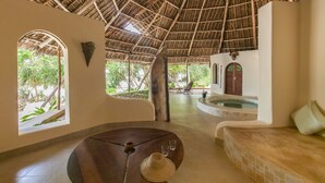 A spacious room with a high thatched ceiling, a round wooden table with a straw hat and glasses, a cushioned bench, and a view of a pool and greenery outside.