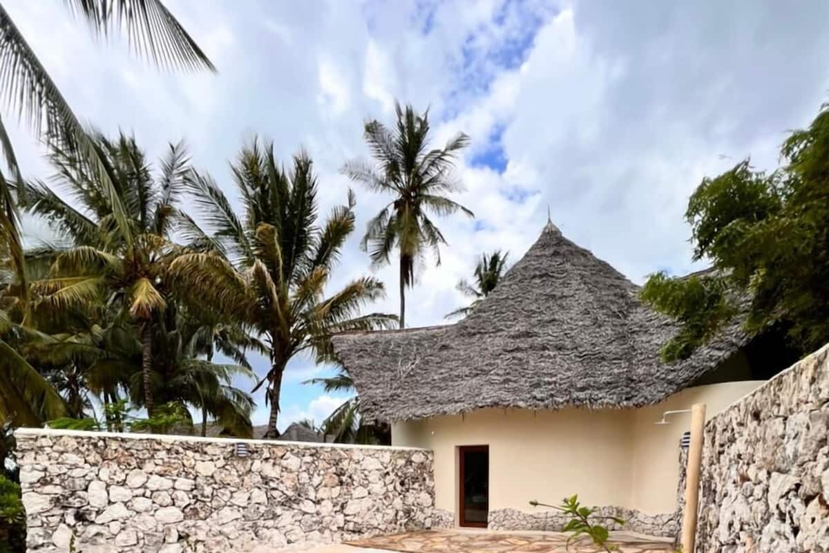 A thatched-roof building with a small pool, surrounded by stone walls and palm trees.