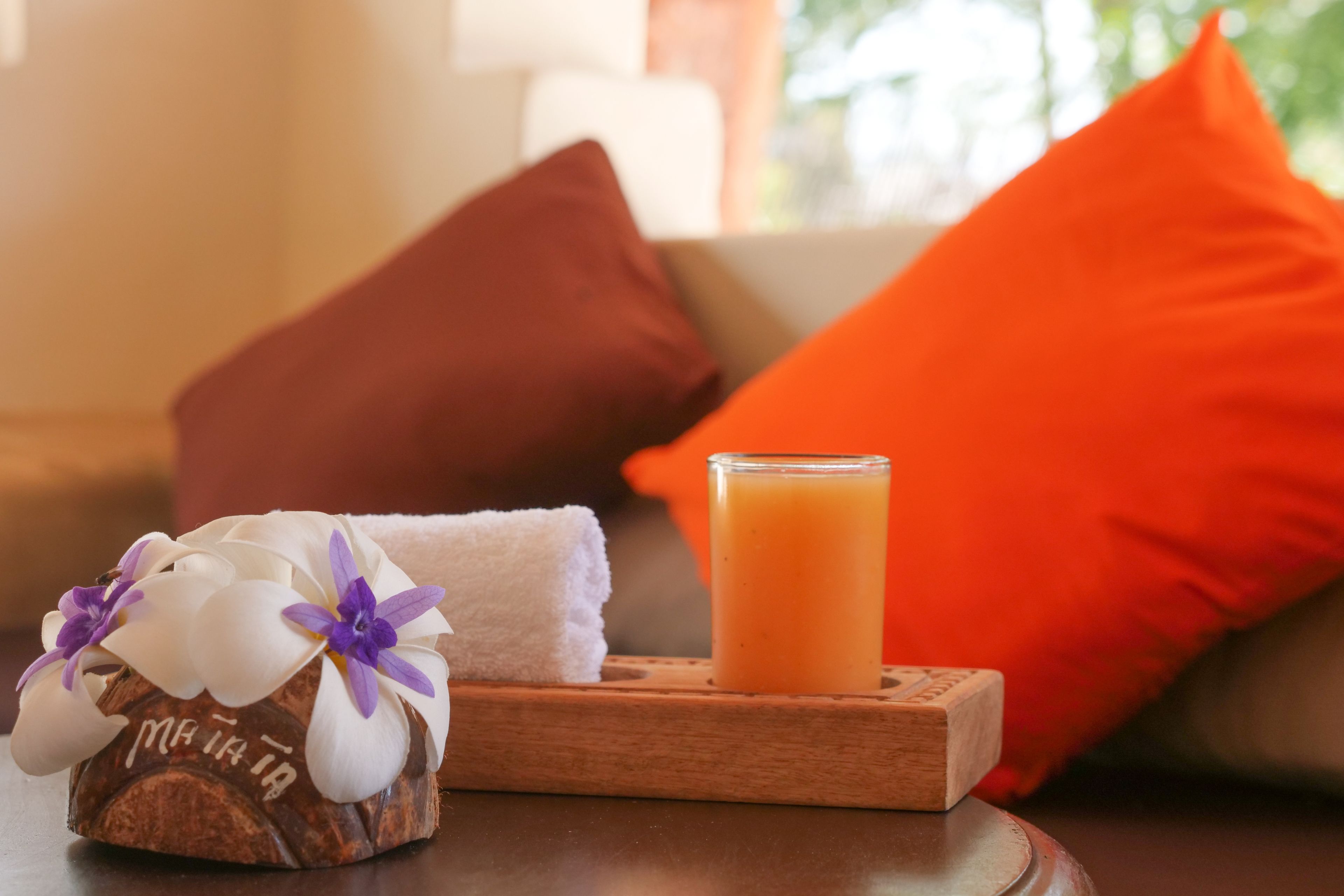 Un plateau en bois avec un verre de jus d’orange, une serviette blanche roulée et une noix de coco décorée de fleurs blanches et violettes.