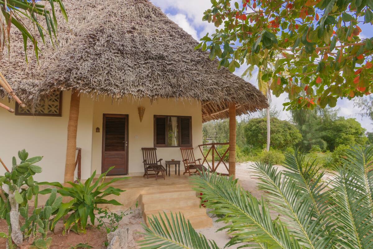 A thatched-roof house with a porch, two wooden chairs, and a small table.