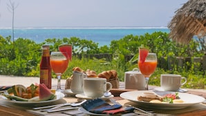 A breakfast table with a view of the ocean, including plates of food, drinks, and a basket of pastries.