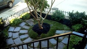 Un chemin de pierres avec un arbre au centre, entouré de pelouse et d’arbustes, et bordé d’une clôture en métal.