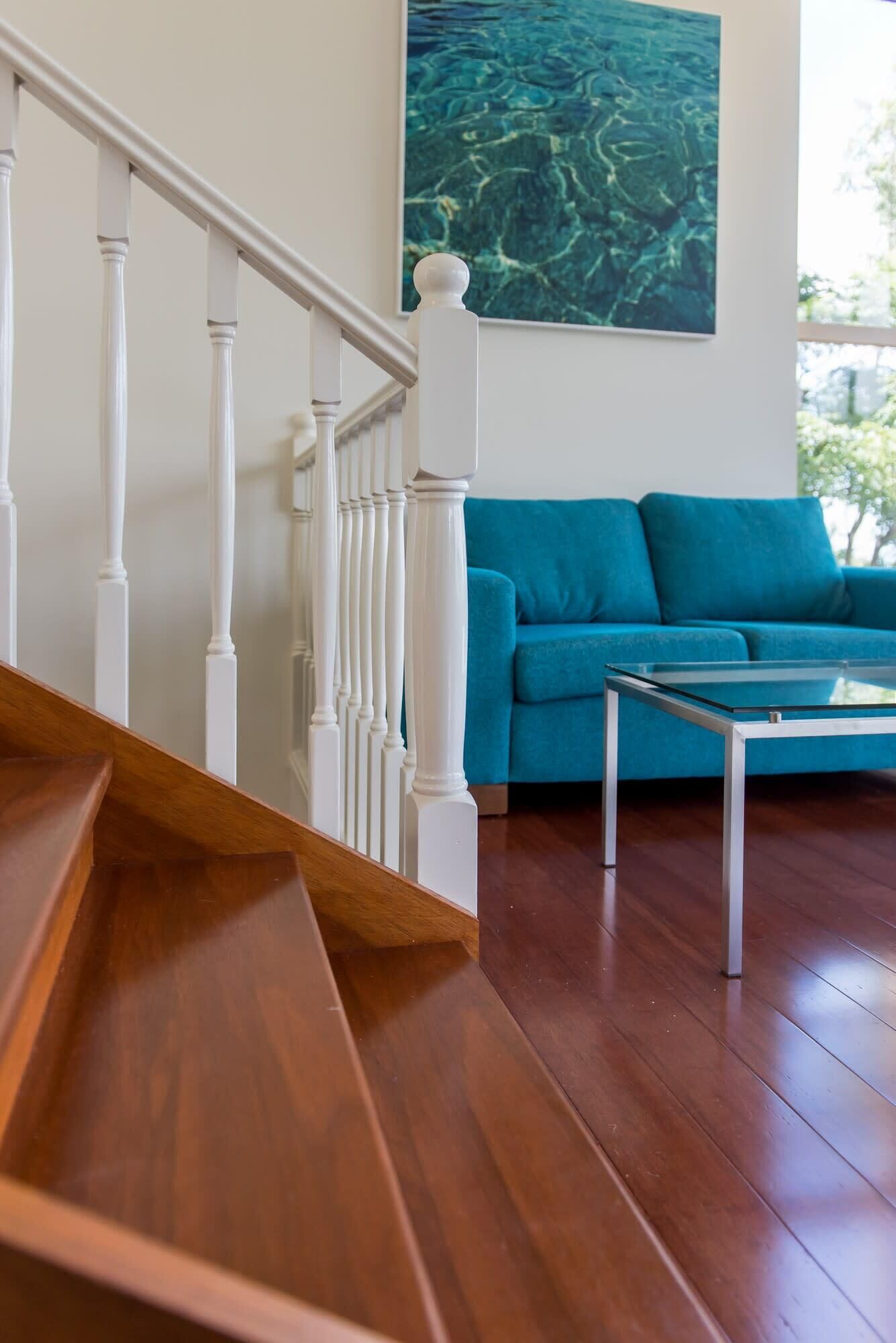 A wooden staircase with white balusters leading to a living room with a teal sofa and a glass coffee table.