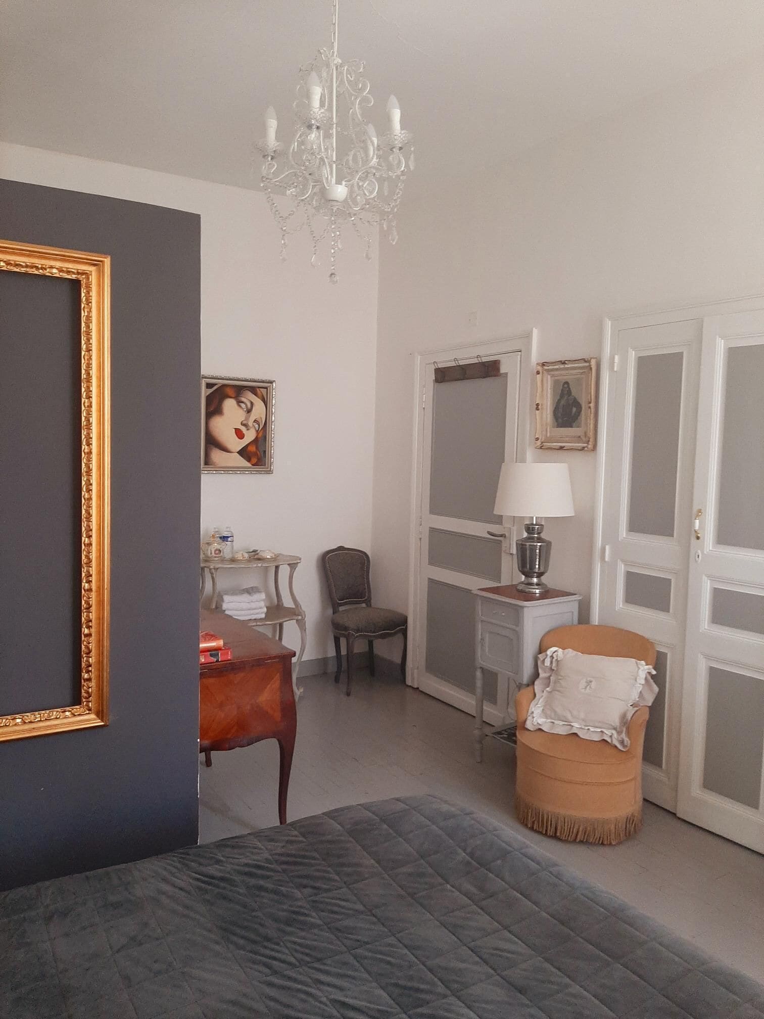 A bedroom with a bed, a chair, a small table, a lamp, and a chandelier.