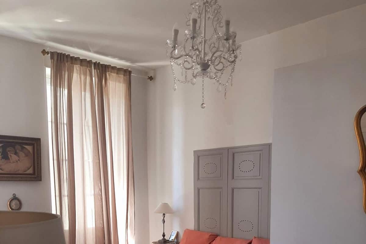 A bedroom with a bed, a nightstand, a dresser, a chandelier, and a window with curtains.
