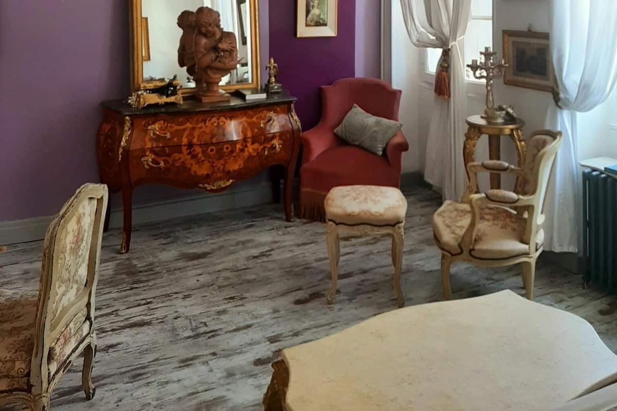 A room with a purple wall, a wooden dresser, a red armchair, a white chair, and a chandelier.