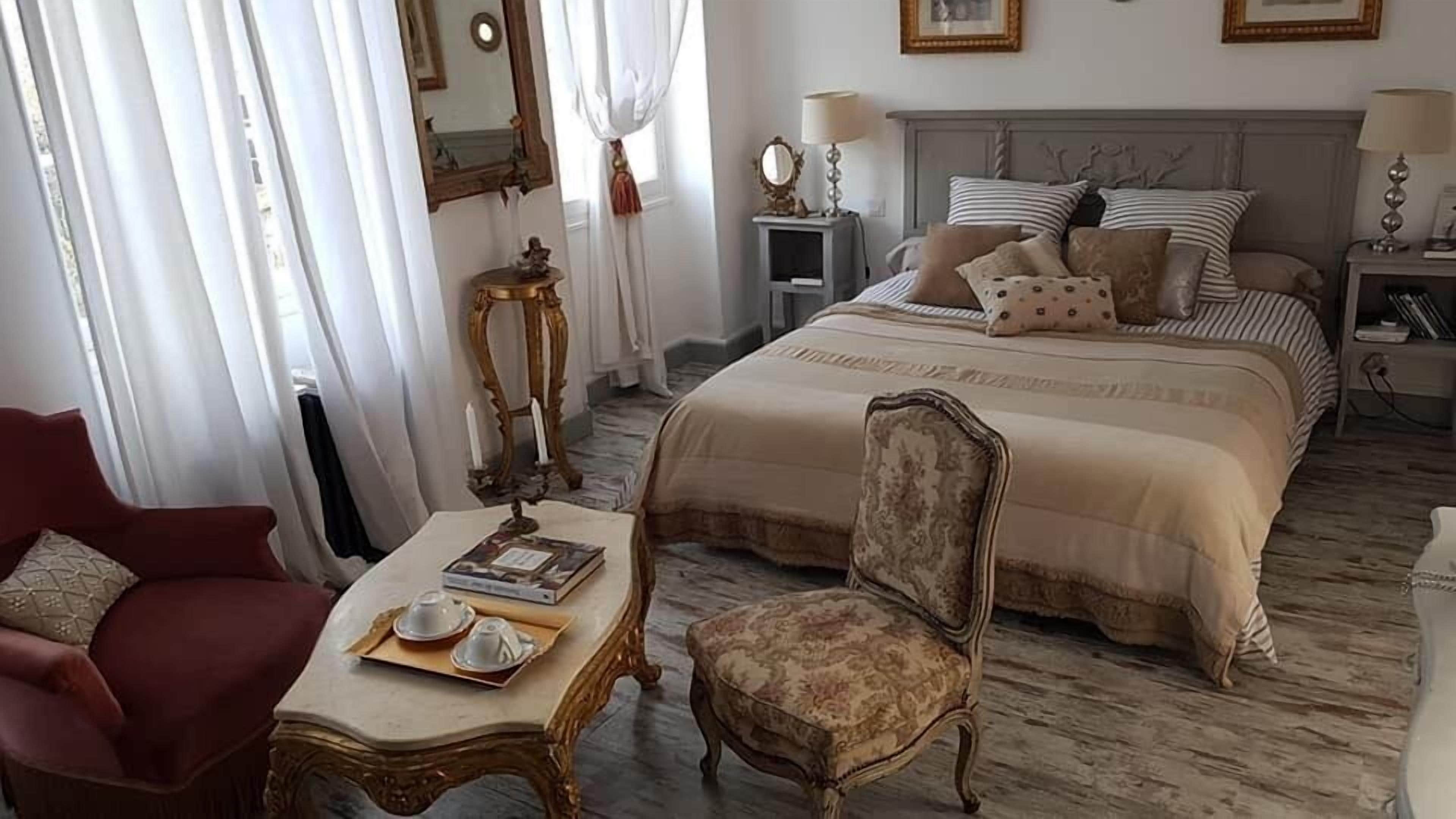 Een slaapkamer met een groot bed, een stoel, een tafel met een boek en theekoppen, en een spiegel aan de muur.