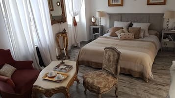 Een slaapkamer met een groot bed, een stoel, een tafel met een boek en theekoppen, en een spiegel aan de muur.