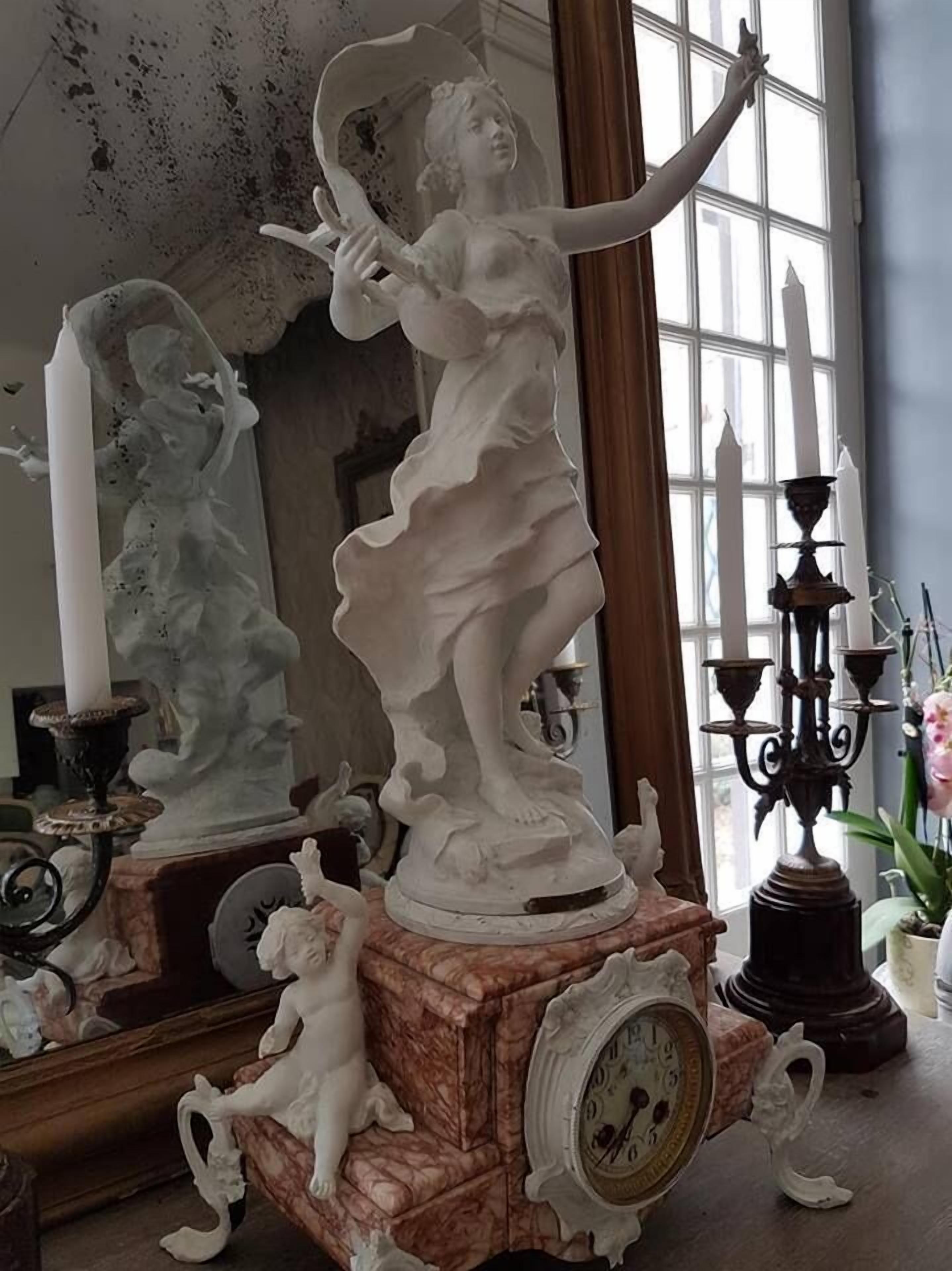 A marble statue on a pedestal with a clock, surrounded by other statues and candelabras in a room with large windows.