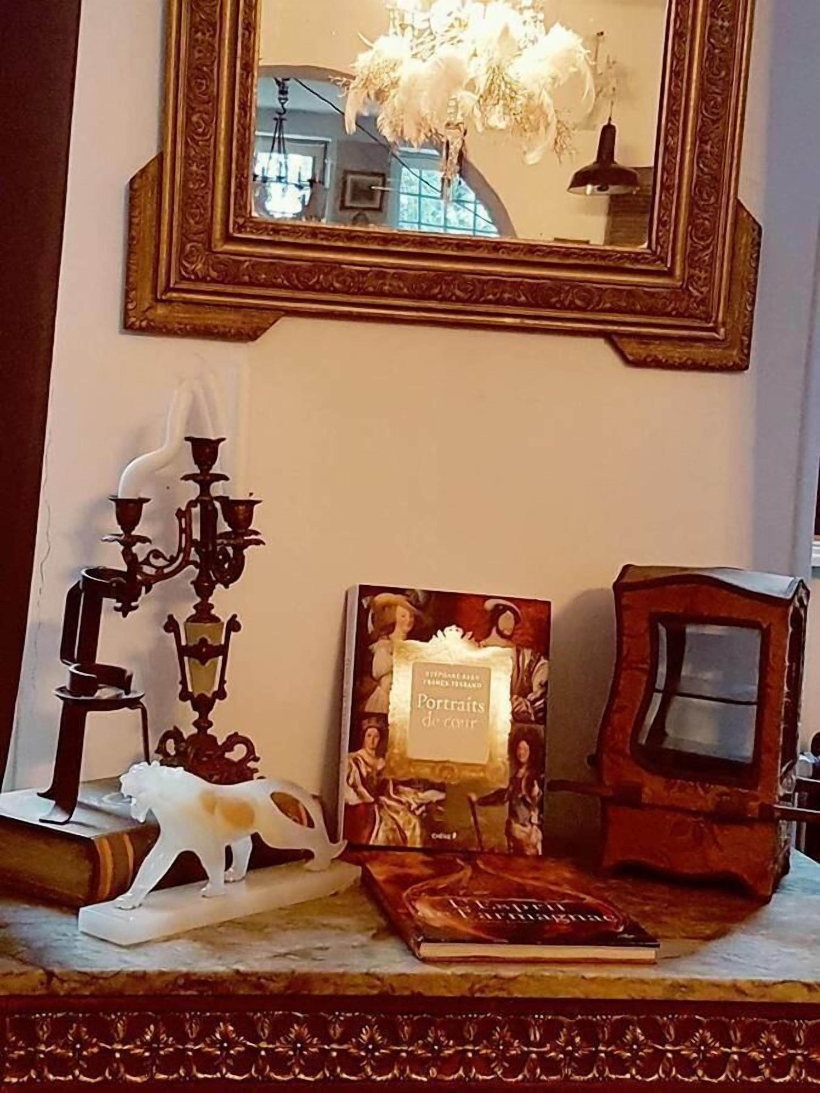 A room with a large ornate mirror, a book titled 'Portraits de Coeur', a decorative candelabra, and a small white figurine on a marble surface.