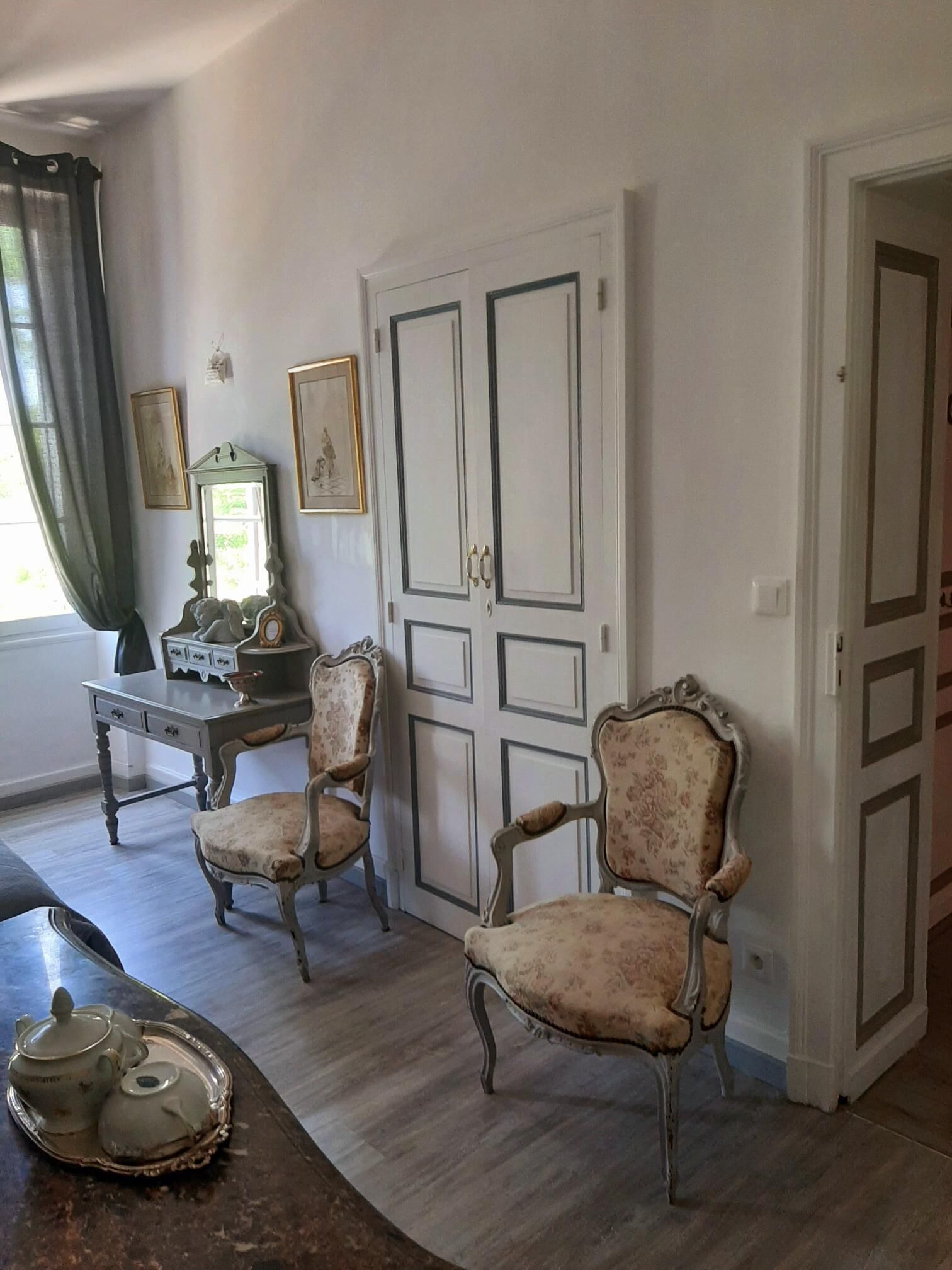 Una habitación con dos sillas decoradas, una mesa con tetera y una puerta con espejo.