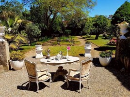 Un espacio al aire libre con mesa redonda y dos sillas, rodeado de plantas en macetas y un sendero en el jardín.