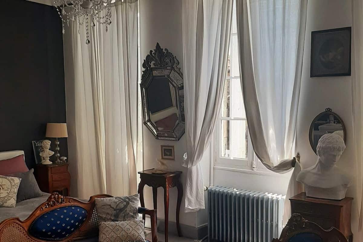 A room with a wooden sofa, a chair, a large mirror, and a bust on a pedestal.