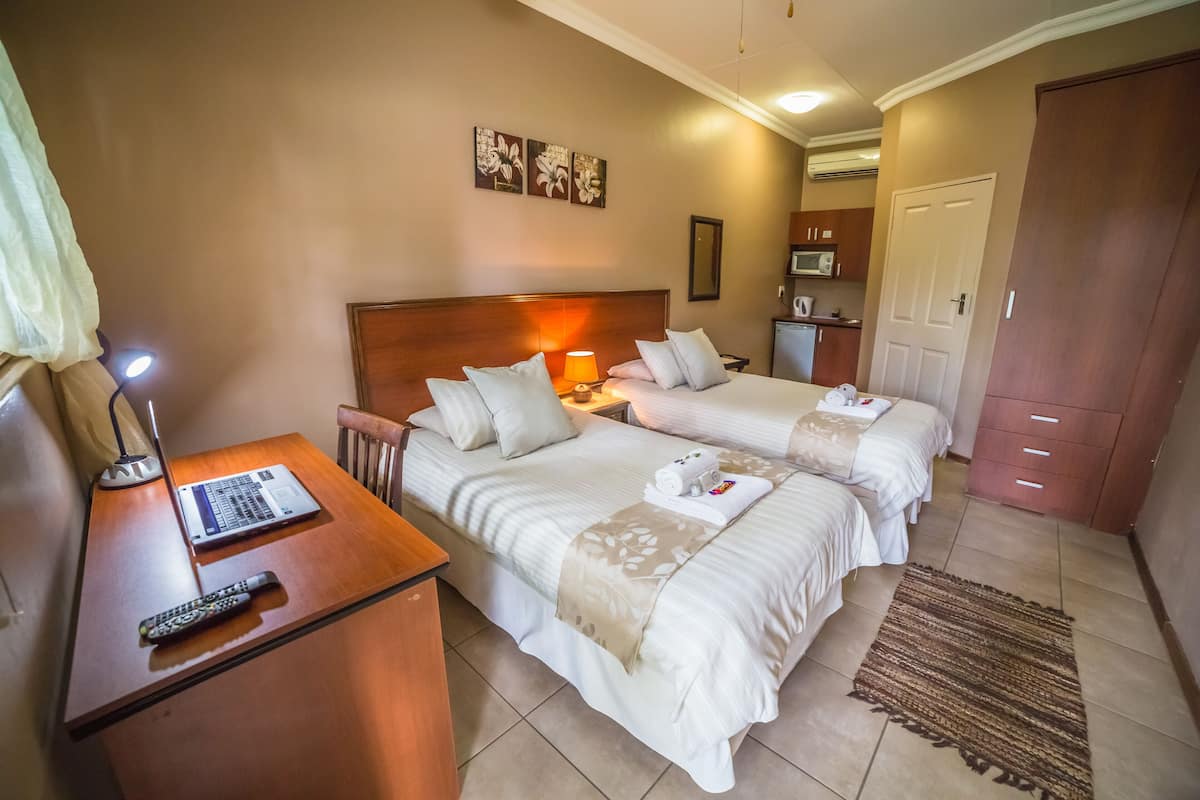 A hotel room with two beds, a desk with a laptop and remote control, a nightstand with a lamp, and a small kitchenette in the background.