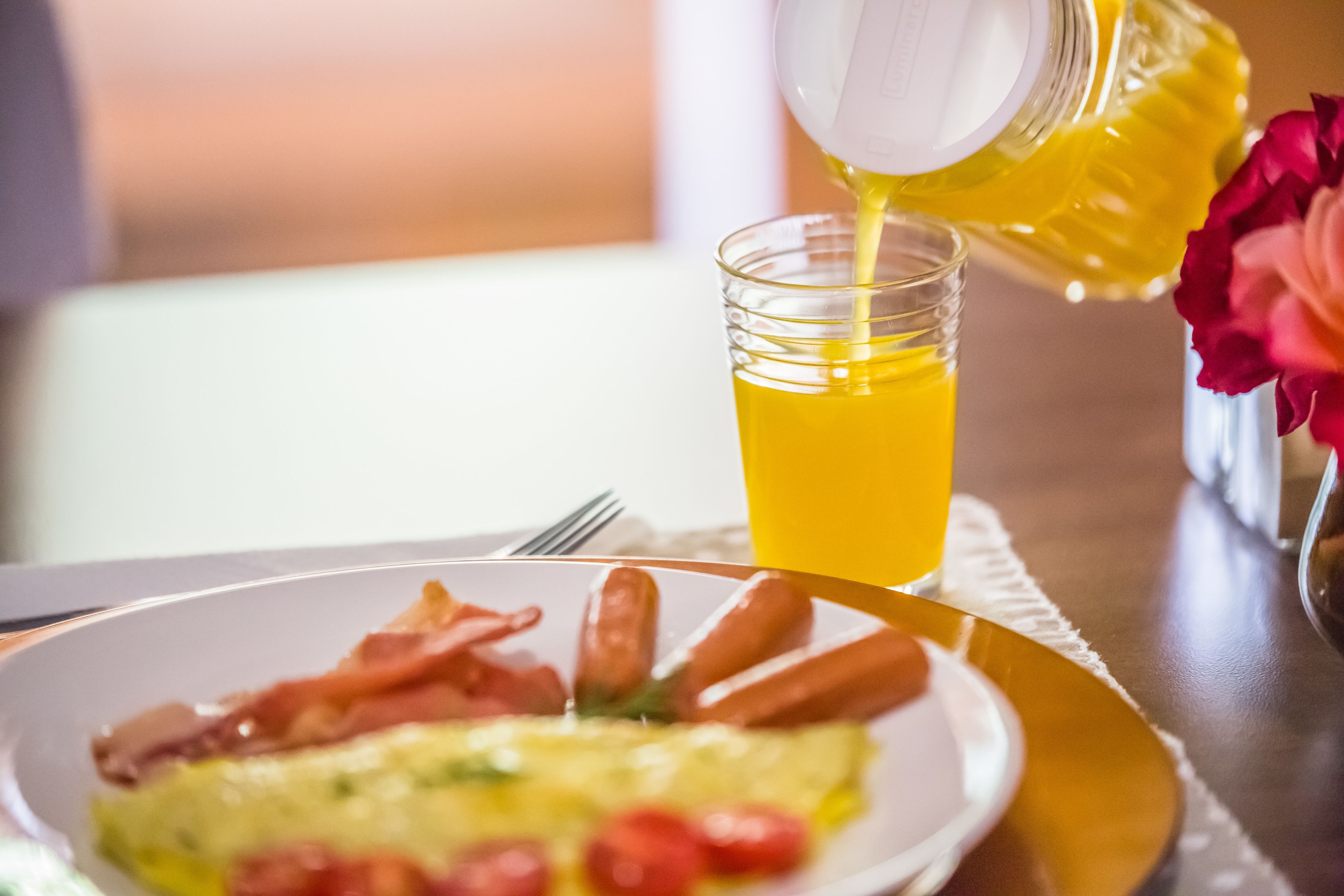 A breakfast setting with a plate of bacon and sausages, a glass of orange juice, and a bottle of orange juice being poured.