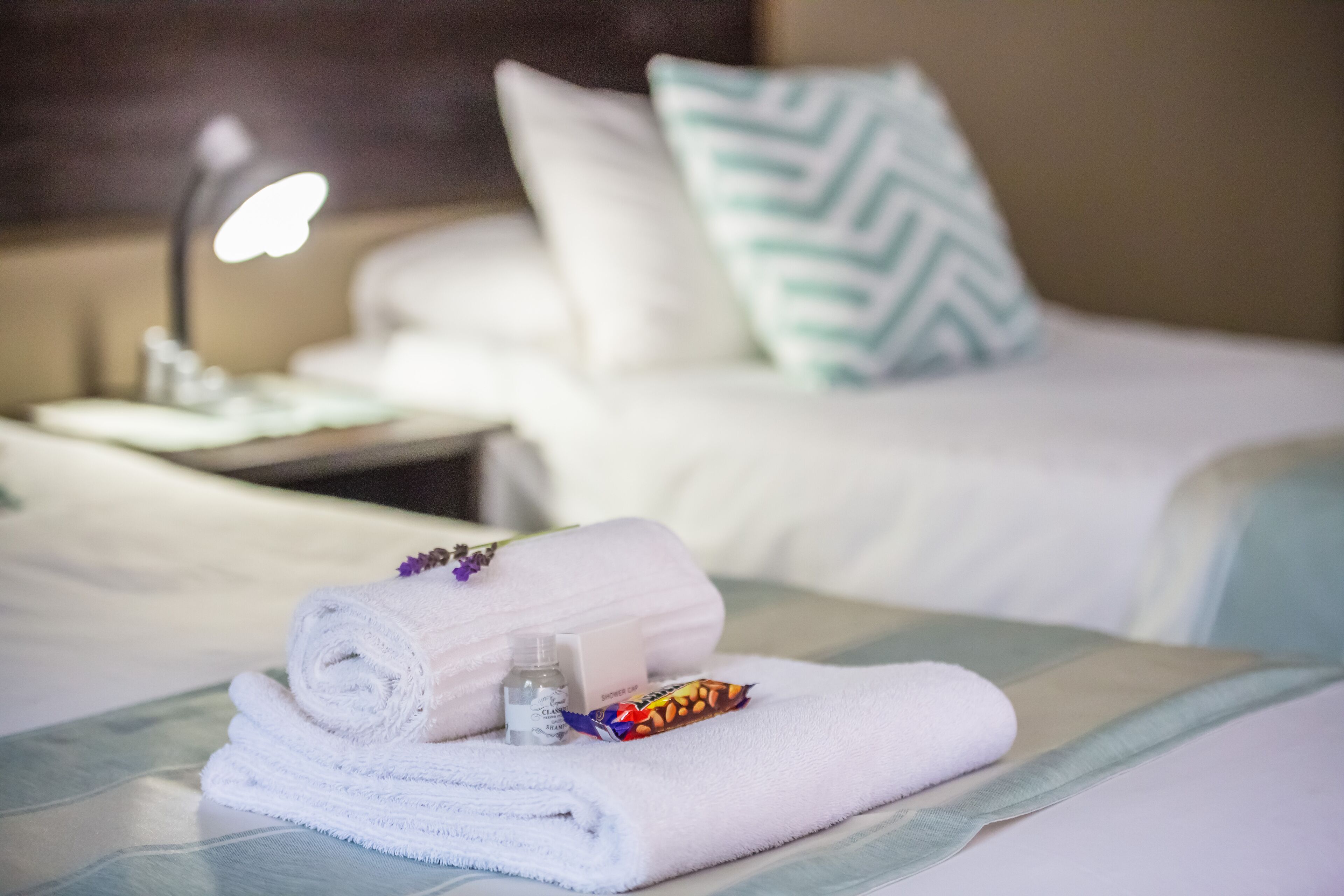 A neatly made bed with a rolled white towel, a small bottle, and a colorful item on the bed.