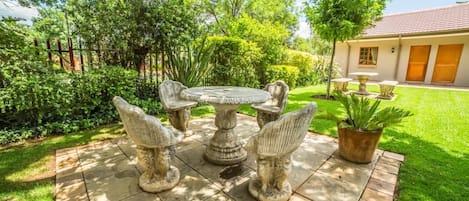 Un patio con una mesa y sillas de piedra, rodeado de abundante vegetación y una casa al fondo.