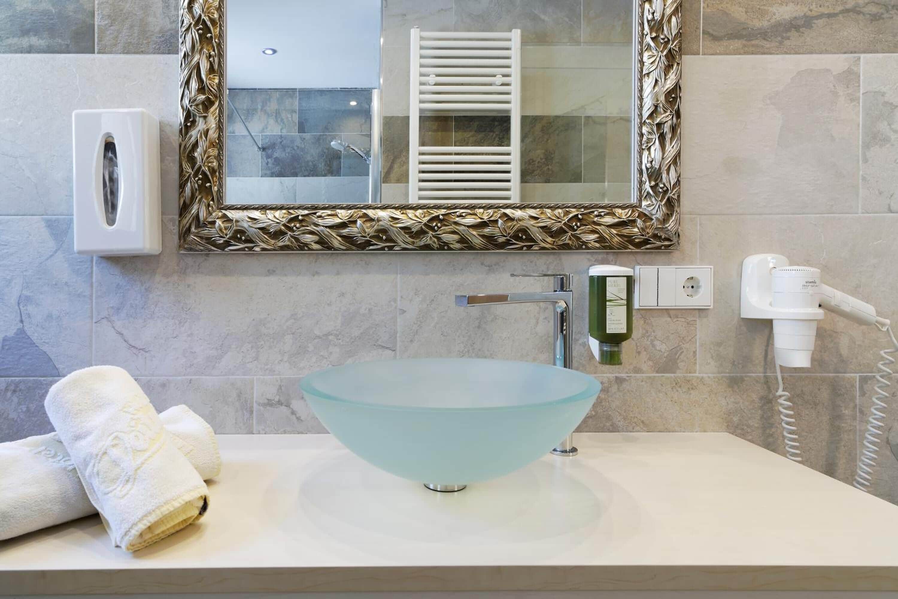 Un bagno con un lavandino in vetro a forma di ciotola, un asciugamano, uno specchio con cornice decorativa e un portascopevoli.