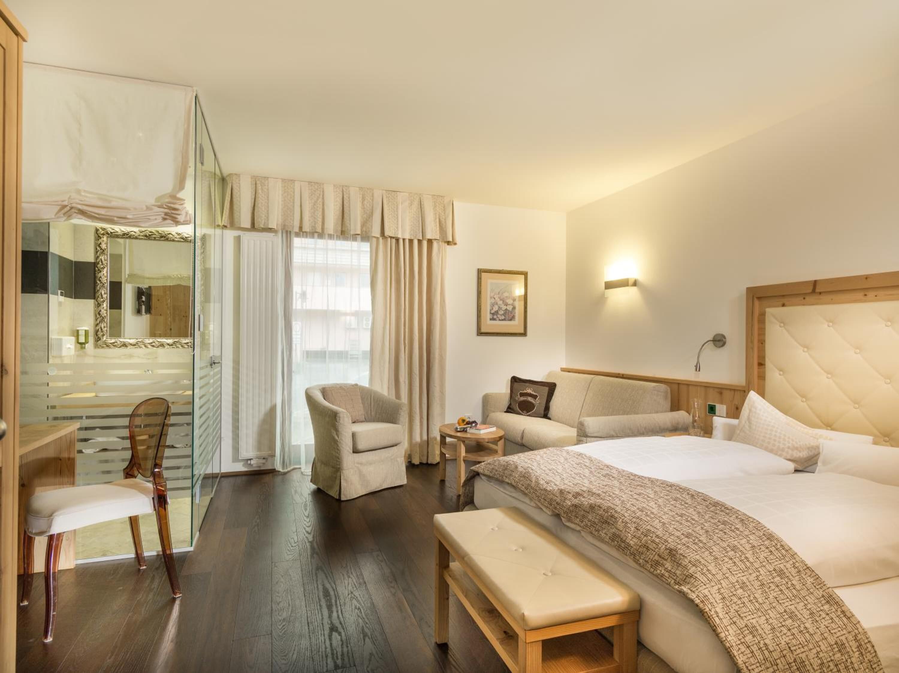 Habitación de hotel con cama, una silla, un banco, una mesita, un sofá, un espejo y una ventana con cortinas.