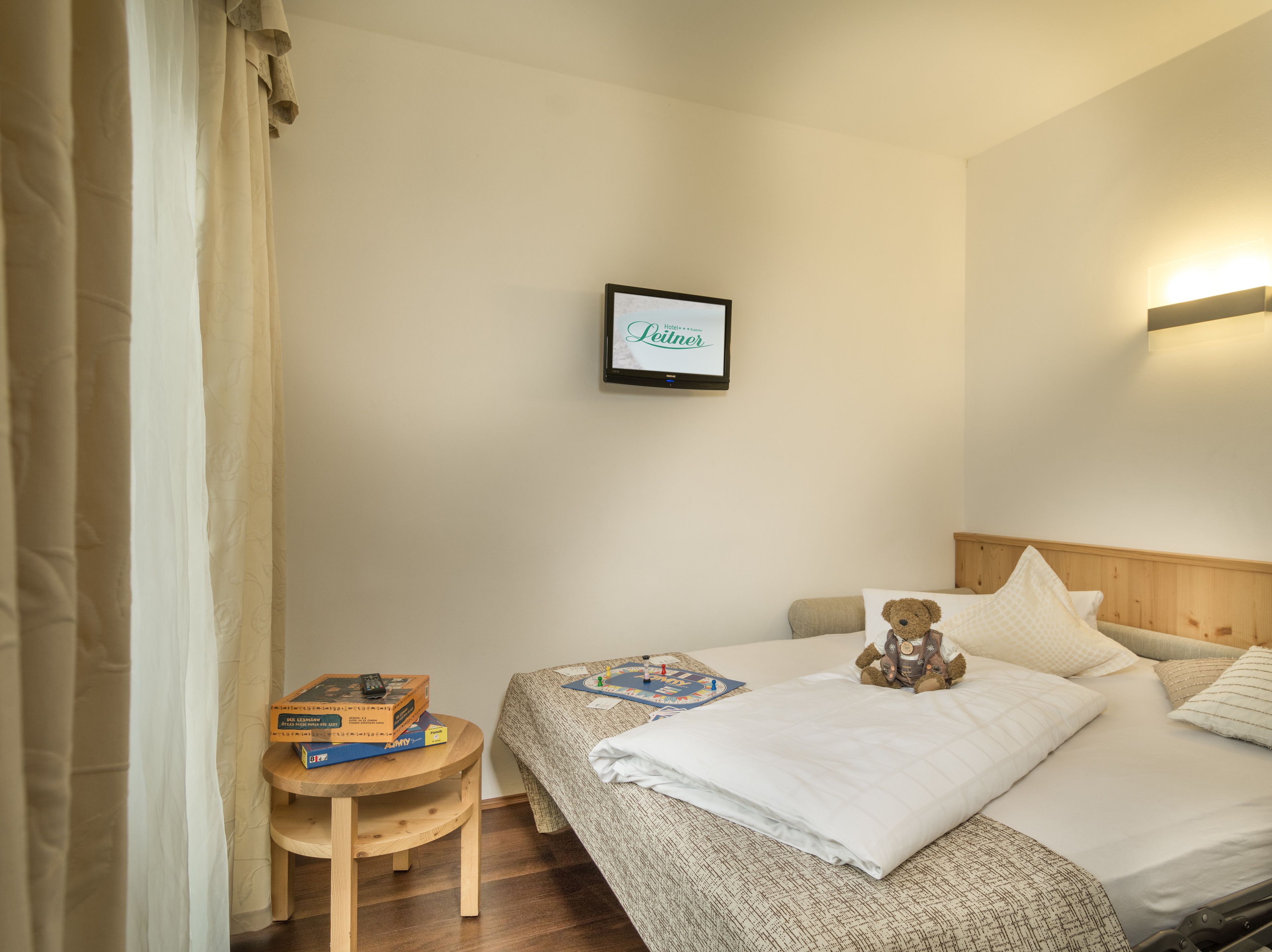 Habitación de hotel con una cama, una pequeña mesa con un juego de mesa, un osito de peluche y un cartel enmarcado en la pared.