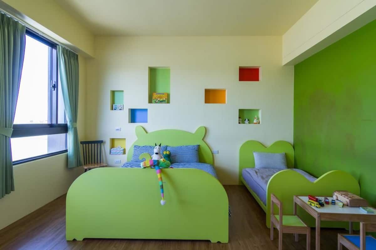 Un dormitorio infantil con una cama verde, una mesita pequeña y una ventana con cortinas.