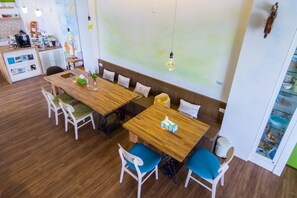 A modern dining area with wooden tables and chairs, a kitchen in the background, and a light fixture above the tables. - Cest Mignon (Dongshan)