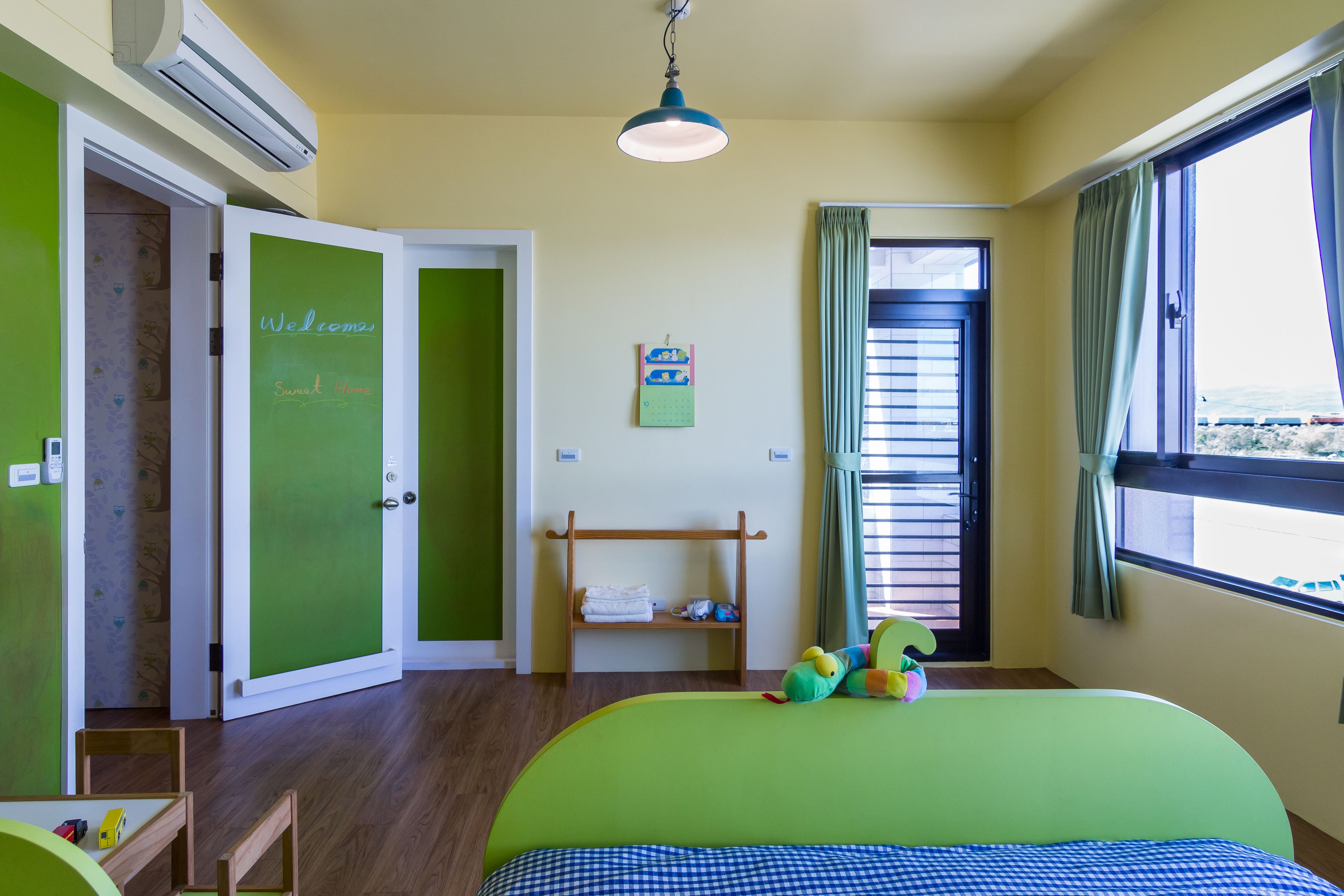 A room with a green bed, a wooden shelf with towels, a window with curtains, and an air conditioning unit.