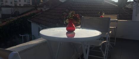 Une table ronde, un vase rouge et des chaises sont disposés sur un balcon donnant sur un quartier résidentiel avec des maisons et des arbres.