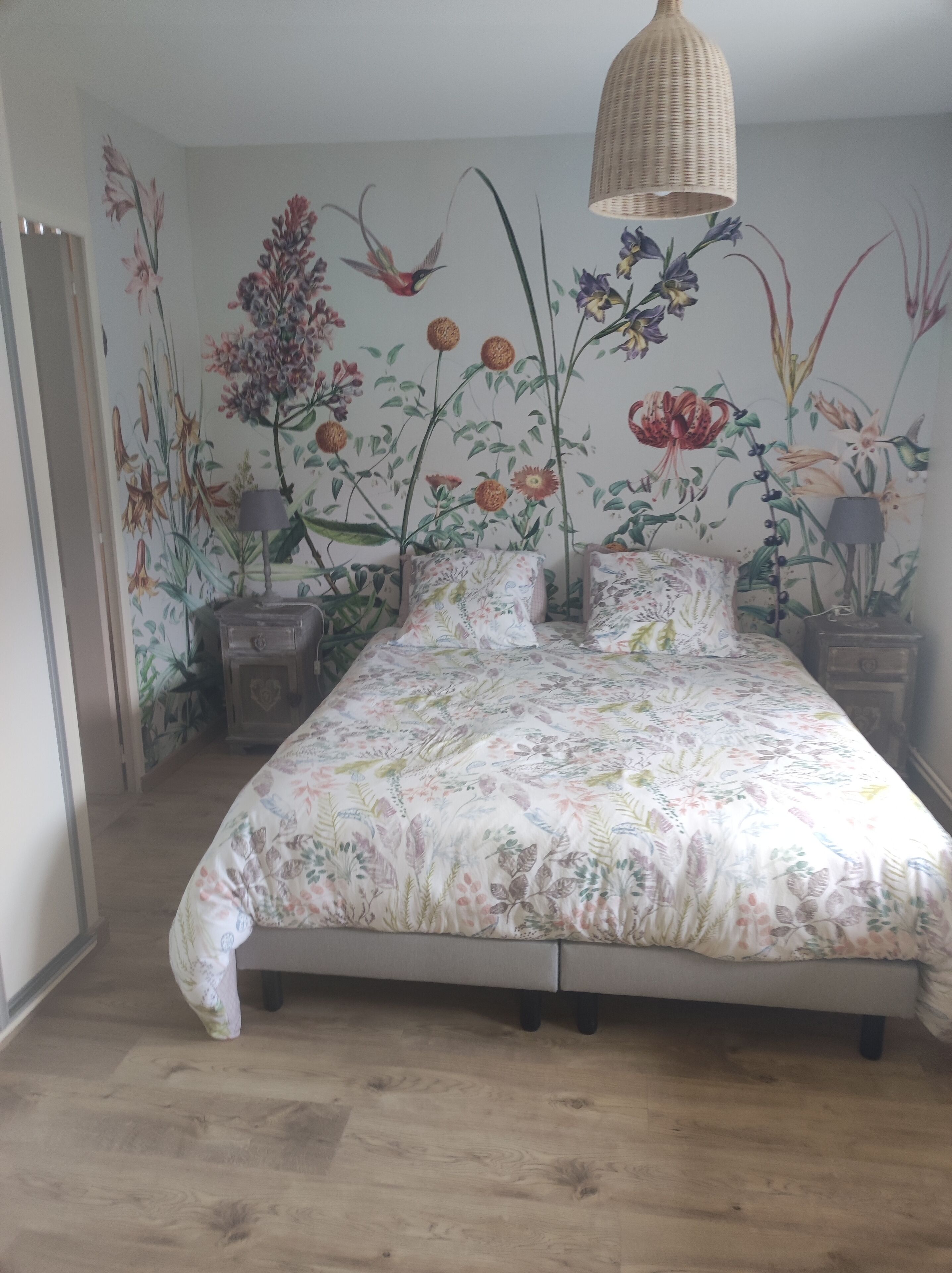 Ein Schlafzimmer mit einem floralen Bettbezug, einer hängenden gewebten Leuchte und einer Wandmalerei mit Blumen und Vögeln.