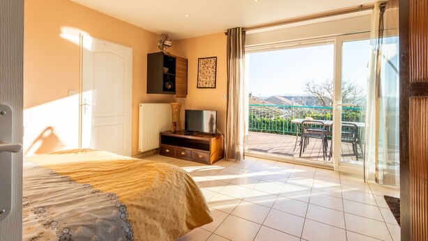 A bedroom with a bed, a TV on a wooden stand, a radiator, and a sliding glass door leading to an outdoor area with a table and chairs.