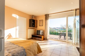 Camera da letto con un letto, una televisione su un mobile in legno, un termosifone e una porta scorrevole in vetro che dà su una zona esterna con tavolo e sedie.