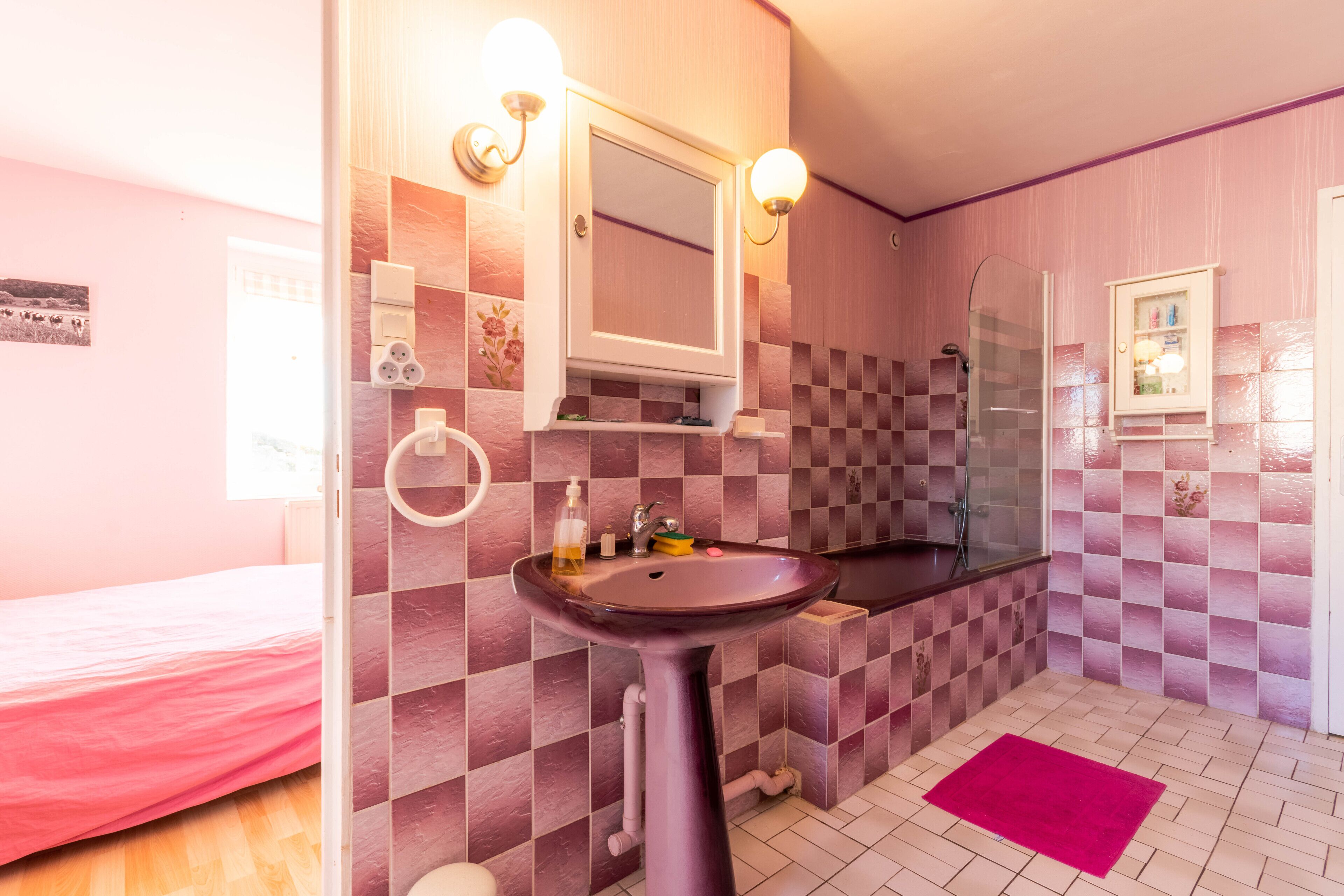 Un baño con pared de baldosas a cuadros, un lavamanos con espejo, una ducha y una cama con colcha rosa.