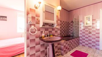 Un baño con pared de baldosas a cuadros, un lavamanos con espejo, una ducha y una cama con colcha rosa.