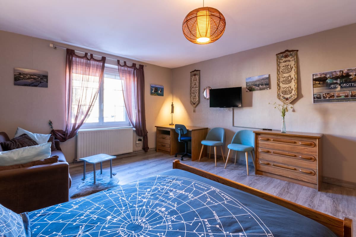 A bedroom with a bed, a desk, a chair, a dresser, a television, and a window with curtains.