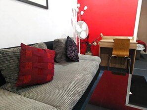 A modern living room with a sofa, a small table, a chair, a fan, and a red accent wall. - Wellesley Hotel (Ilford)