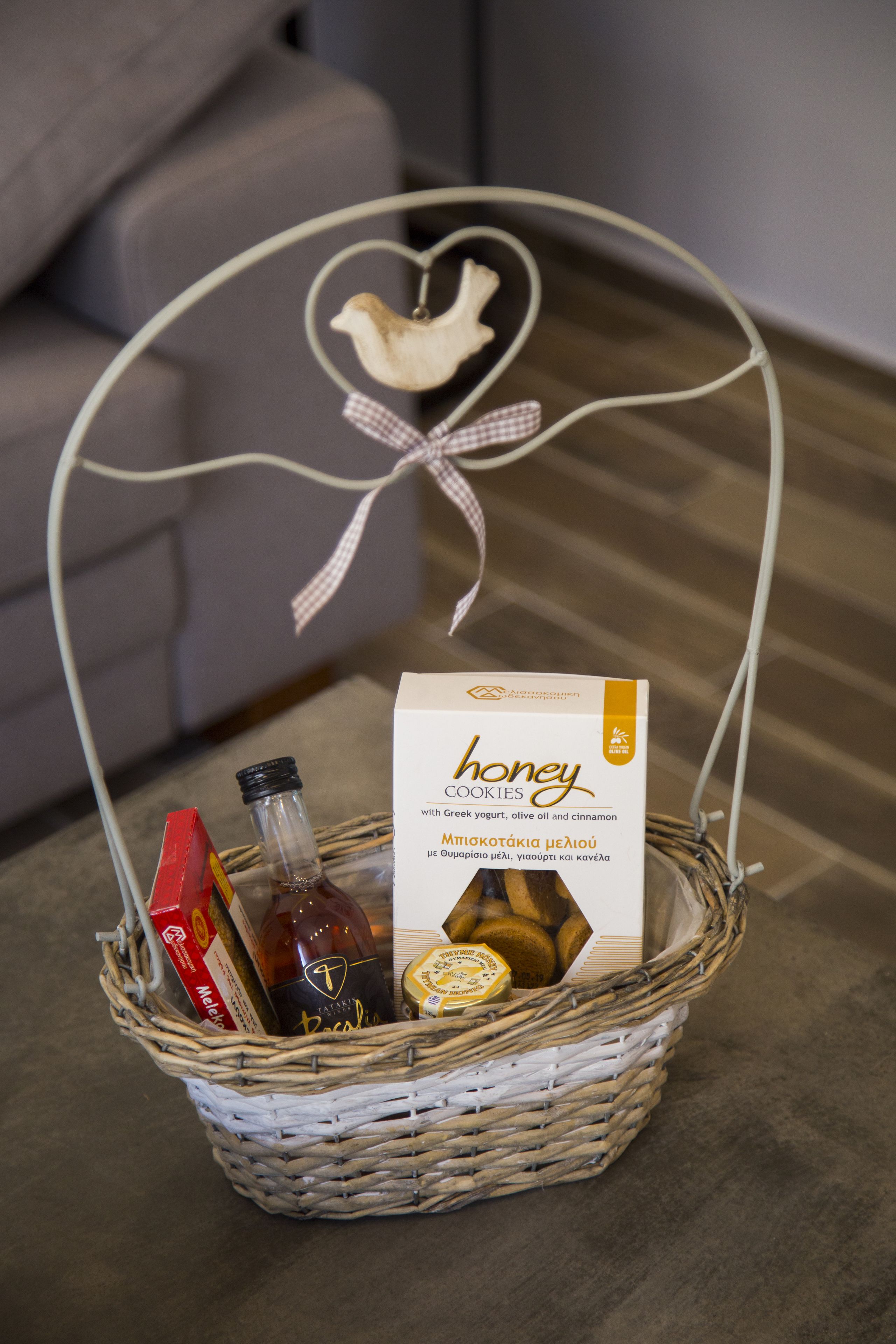 A wicker basket with a heart-shaped handle, containing a box of honey cookies, a bottle of olive oil, and a bottle of red wine.