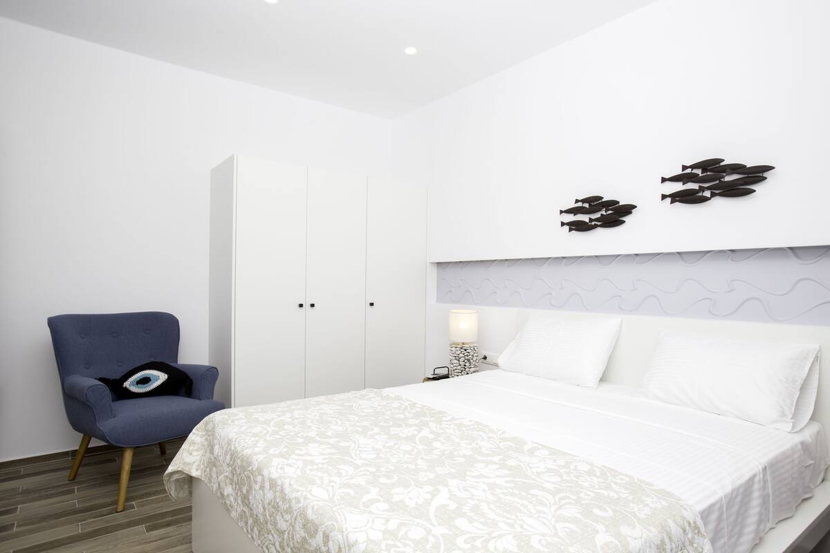 A bedroom with a bed, a chair, a nightstand, and a wall-mounted light fixture.