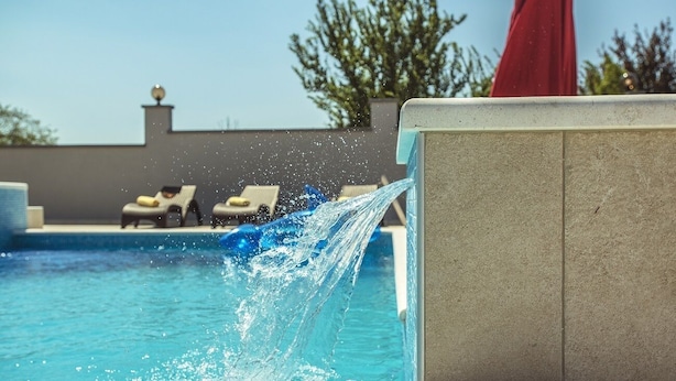 Una piscina con agua cayendo por una pared, sillas de descanso y un paraguas rojo.