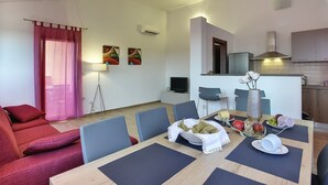 Una sala de estar moderna con un sofá rojo, una mesa de comedor dispuesta con frutas y un florero, y una cocina al fondo.