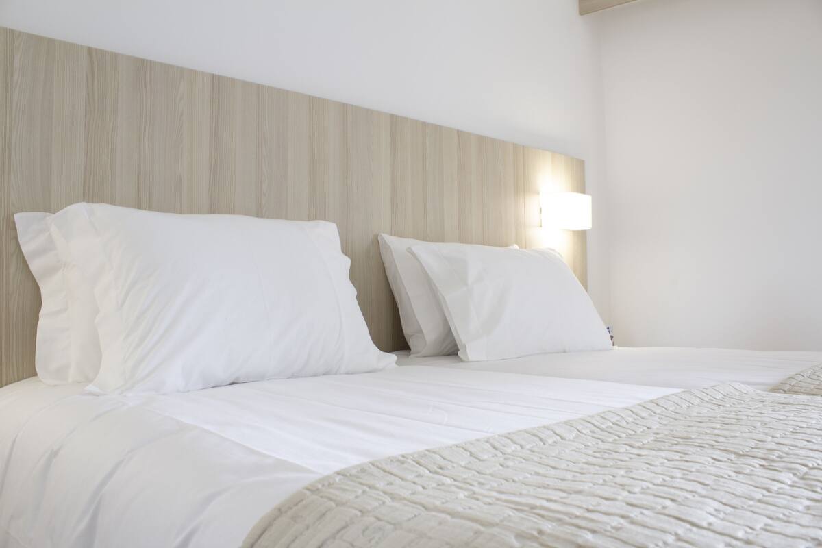 A hotel room with a wooden headboard, two white beds with pillows, and a bedside lamp.